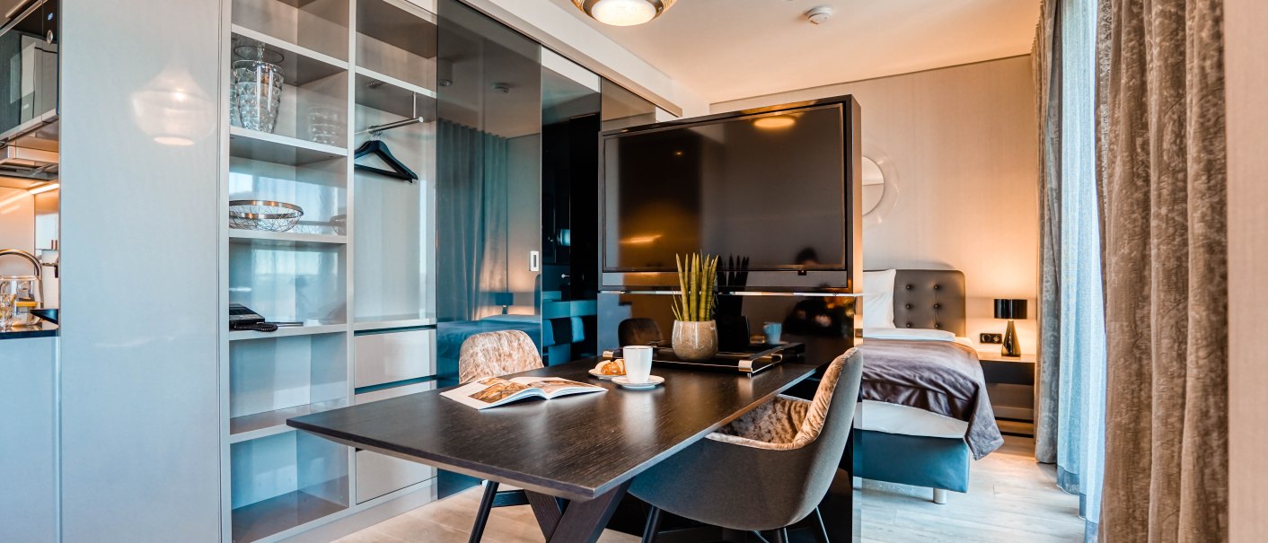 Moderne Suite mit Esstisch, Stühlen, großem Fernseher und Bett. Helle Holzböden, stilvolle Beleuchtung und Vorhänge schaffen eine elegante Atmosphäre., © CLOUD N°7 APARTMENTS GmbH