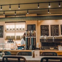 Modernes Café mit Theke, Kaffeemaschine und Speisekarten an der Wand. Pflanzen im Vordergrund, gemütliche Atmosphäre., © Maison Karl, Stuttgart Modernes Café mit Theke, Kaffeemaschine und Speisekarten an der Wand. Pflanzen im Vordergrund, gemütliche Atmosphäre., © Maison Karl, Stuttgart