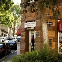 Entrance to the Fr&ouml;hlich coffee roastery on a street corner with a red awning and signs. Cars are parked along the street, trees provide shade., &copy; Kaffeer&ouml;sterei Fr&ouml;hlich