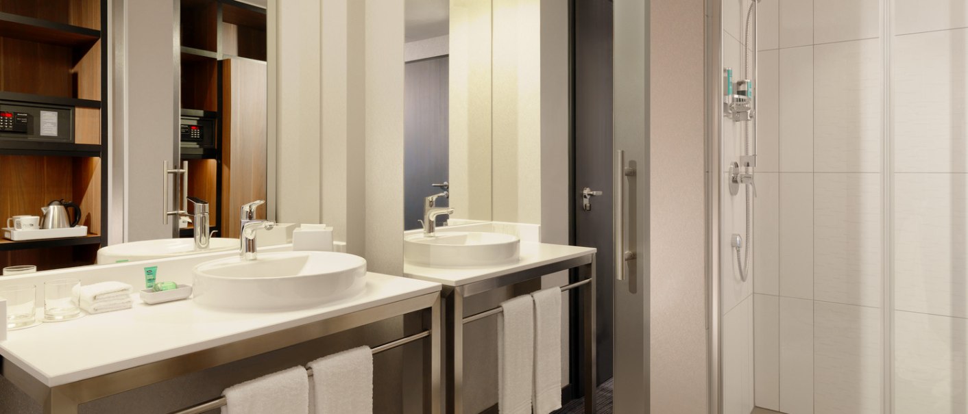 Ein modernes Badezimmer mit zwei Waschbecken, gro&szlig;en Spiegeln und einer Dusche mit Glast&uuml;r. Handt&uuml;cher und Pflegeprodukte sind bereitgestellt., &copy; Aloft Stuttgart