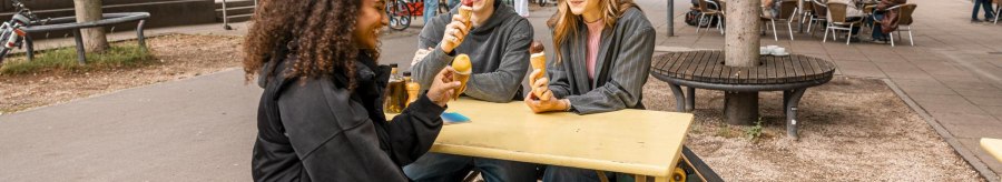 Drei Personen sitzen an einem gelben Tisch im Freien und essen Eis. Im Hintergrund sind B&auml;ume, Fahrr&auml;der und ein Caf&eacute; zu sehen., &copy; Stuttgart Marketing GmbH, Sarah Schmid