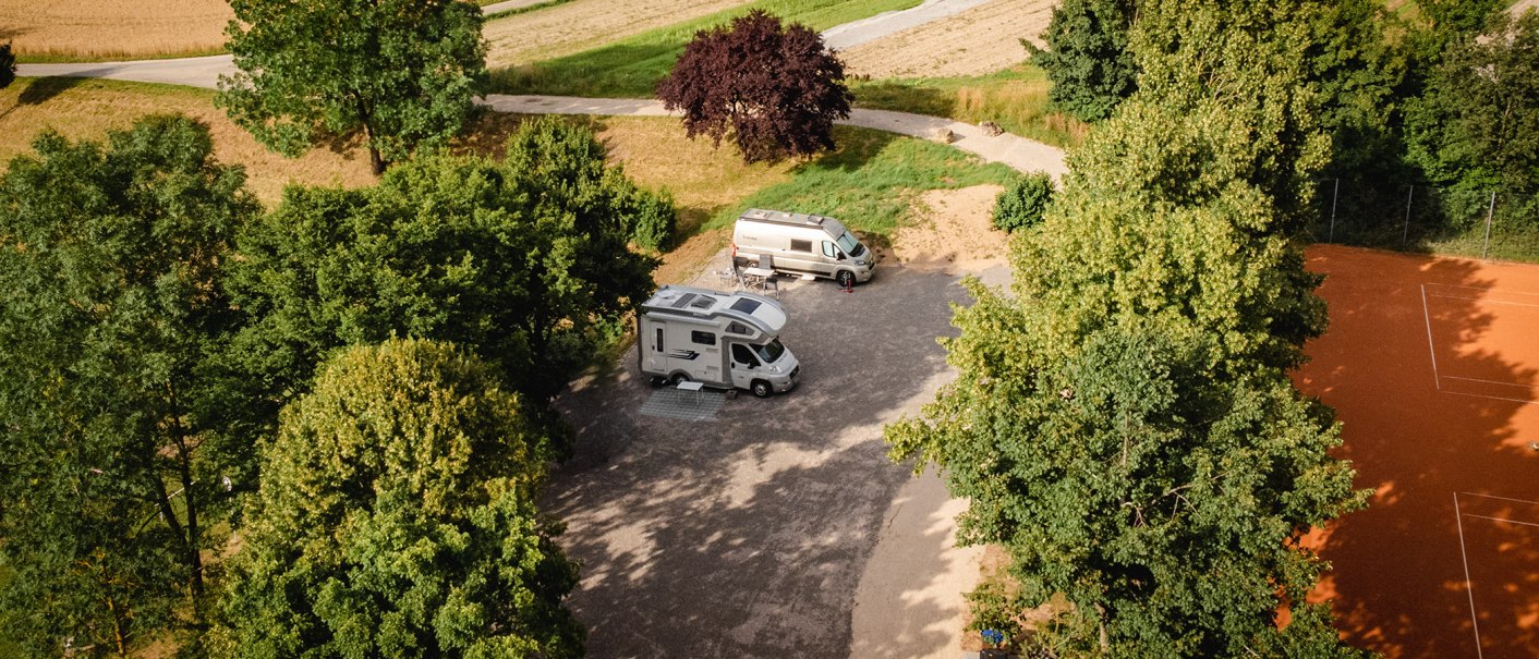 Two mobile homes are parked on a pitch surrounded by trees. A tennis court can be seen on the right of the picture., © Blu Dolci Artwork Two mobile homes are parked on a pitch surrounded by trees. A tennis court can be seen on the right of the picture., © Blu Dolci Artwork
