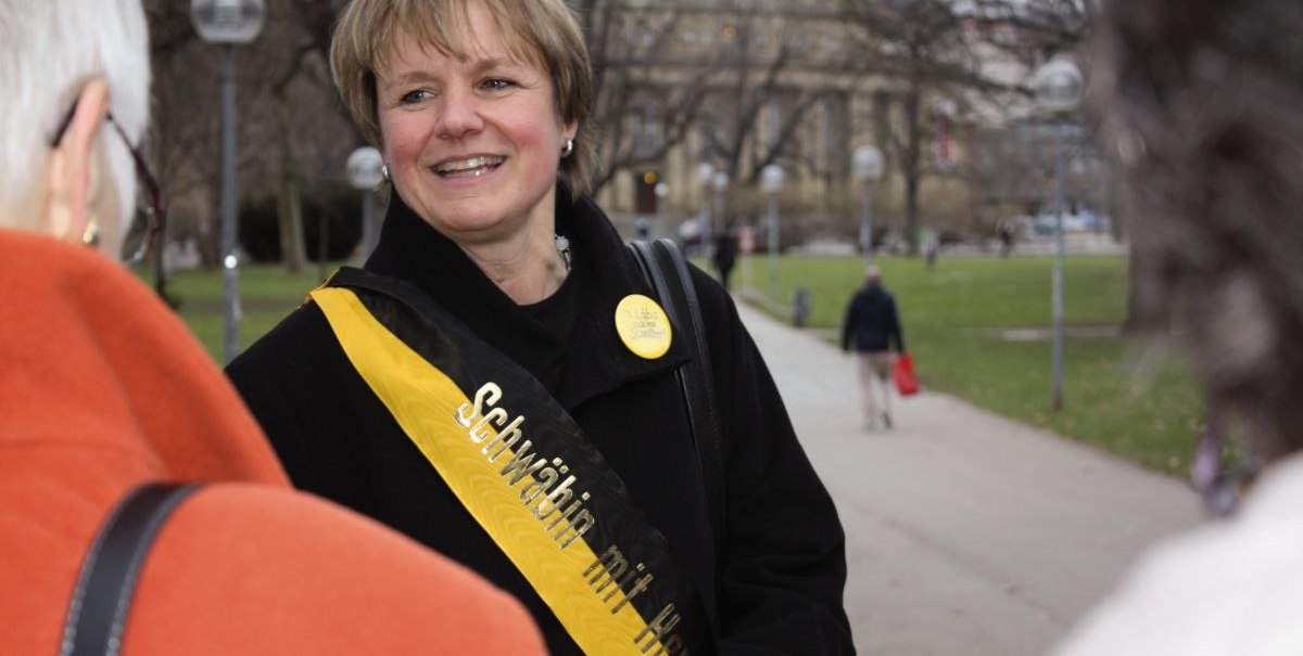 Eine Frau mit einer Schärpe 'Schwäbin mit Herz' lächelt und spricht mit anderen Personen in einem Park., © Stuttgart-Marketing GmbH Eine Frau mit einer Schärpe 'Schwäbin mit Herz' lächelt und spricht mit anderen Personen in einem Park., © Stuttgart-Marketing GmbH