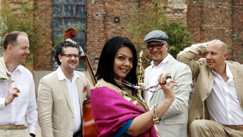 Eine Frau in einem bunten Kleid posiert mit vier Männern in hellen Anzügen vor einer alten Backsteinmauer. Sie lächeln und wirken entspannt., © Round midnight music Eine Frau in einem bunten Kleid posiert mit vier Männern in hellen Anzügen vor einer alten Backsteinmauer. Sie lächeln und wirken entspannt., © Round midnight music