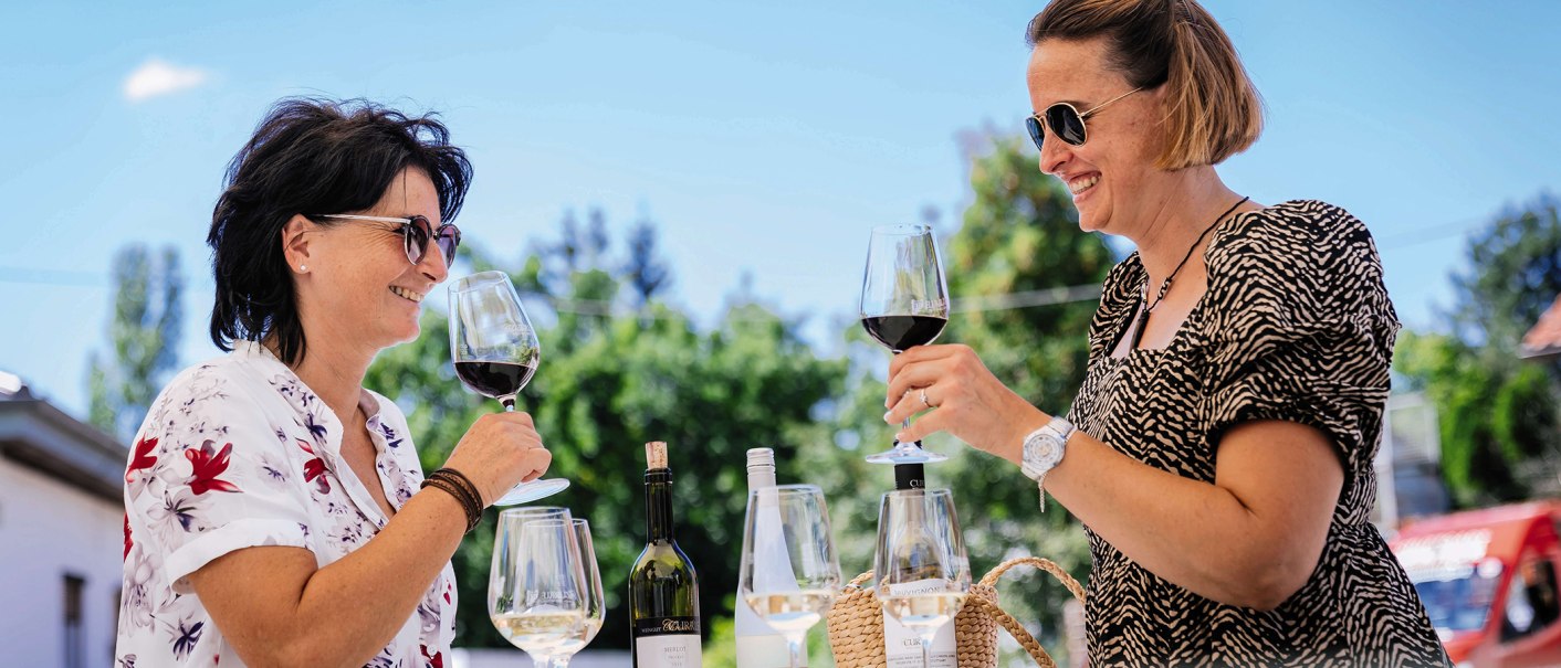 Zwei Frauen genießen Wein im Freien, stoßen mit Gläsern an. Im Hintergrund sind Bäume und ein blauer Himmel zu sehen., © SMG, Thomas Niedermüller