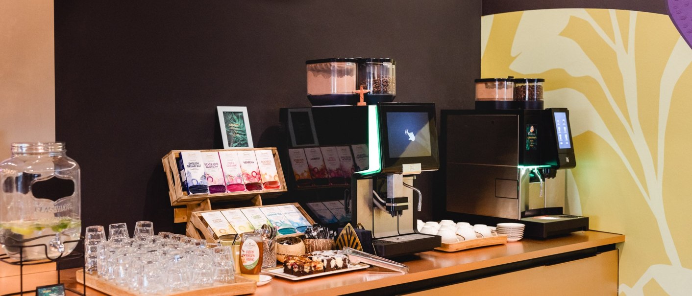 Selbstbedienungsbereich mit Kaffeemaschine, Teesortiment, Gläsern und Snacks auf einem Tresen., © TOMAS