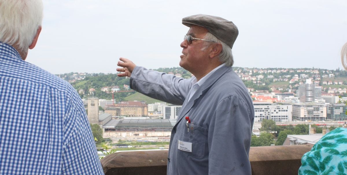 Ein älterer Mann in Arbeitskleidung und Mütze zeigt auf eine Stadtlandschaft. Im Hintergrund sind Gebäude und Hügel zu sehen., © Stuttgart-Marketing GmbH Ein älterer Mann in Arbeitskleidung und Mütze zeigt auf eine Stadtlandschaft. Im Hintergrund sind Gebäude und Hügel zu sehen., © Stuttgart-Marketing GmbH
