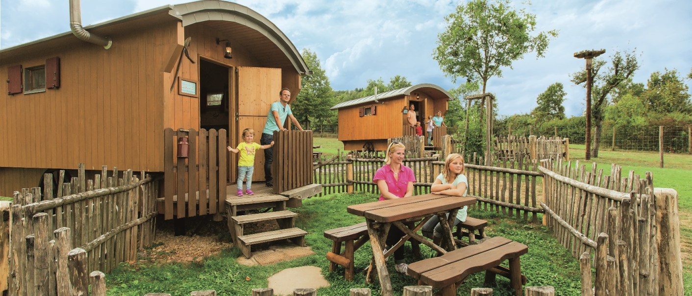 A family in front of a shepherd's wagon at a campground. A wooden fence, a picnic table, and a green meadow. Two shepherd's wagons in the background, blue sky., &copy; Tripsdrill Natur-Resort GmbH