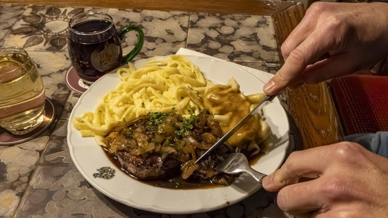 Ein Teller mit Zwiebelrostbraten, Spätzle und Soße. Daneben stehen zwei Gläser mit Getränken. Eine Person schneidet das Fleisch., © Stuttgart-Marketing GmbH, Sarah Schmid