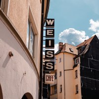 Schild mit 'WEISS Caf&eacute;' an einem Geb&auml;ude, daneben ein weiteres Haus mit Ger&uuml;st. Blauer Himmel mit Wolken im Hintergrund., &copy; SMG_Sarah Schmid