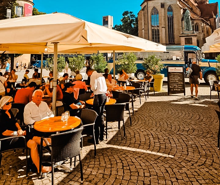 Terrasse am Schillerplatz mit Menschen unter Sonnenschirmen, im Hintergrund eine Kirche und ein Turm bei klarem Himmel., © Alte Kanzlei Stuttgart Terrasse am Schillerplatz mit Menschen unter Sonnenschirmen, im Hintergrund eine Kirche und ein Turm bei klarem Himmel., © Alte Kanzlei Stuttgart