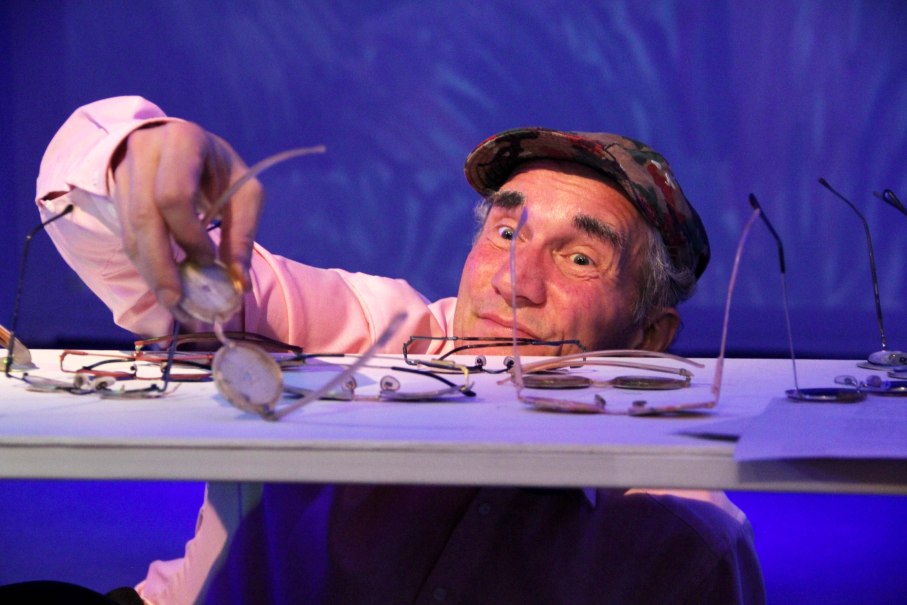 Ein Mann mit einer M&uuml;tze greift l&auml;chelnd nach einer Brille auf einem Tisch, der mit mehreren Brillen bedeckt ist. Der Hintergrund ist blau beleuchtet., &copy; Katja Ritter