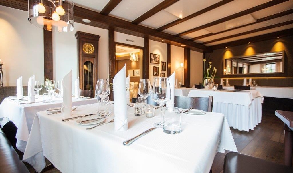 An elegant restaurant with white tablecloths, glasses, and silverware. A ceiling with exposed wooden beams, a wall clock, and a mirror in the background. A cozy, stylish atmosphere., &copy; Hotel Waldhorn