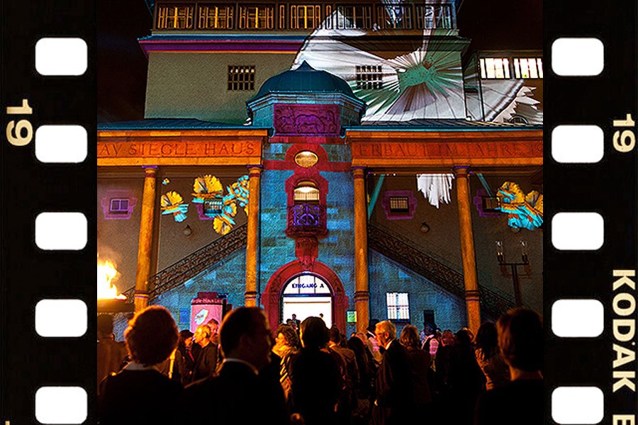 Das Gustav-Siegle-Haus ist nachts bunt beleuchtet. Eine Menschenmenge steht davor. Links und rechts sind Filmstreifen zu sehen., © Stuttgarter Philharmoniker Das Gustav-Siegle-Haus ist nachts bunt beleuchtet. Eine Menschenmenge steht davor. Links und rechts sind Filmstreifen zu sehen., © Stuttgarter Philharmoniker