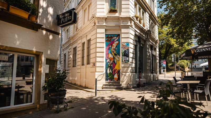 Eine Stra&szlig;enecke mit einem Pub-Schild, bunter Wandkunst und Caf&eacute;-Tischen unter einem Sonnenschirm., &copy; SMG Stuttgart Marketing GmbH - Sarah Schmid