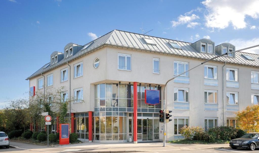 Modernes Hotelgebäude mit Glasfassade und roten Säulen. Mehrere Fenster, blauer Himmel, Autos und Ampel im Vordergrund., © Hotel Stuttgart-Sindelfingen City by Tulip Inn Modernes Hotelgebäude mit Glasfassade und roten Säulen. Mehrere Fenster, blauer Himmel, Autos und Ampel im Vordergrund., © Hotel Stuttgart-Sindelfingen City by Tulip Inn