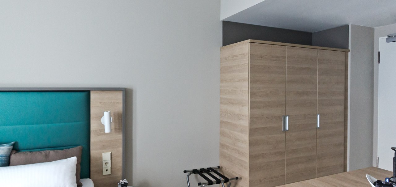 Ein modernes Schlafzimmer mit einem gro&szlig;en Holzschrank, einem Bett mit t&uuml;rkisfarbenem Kopfteil und einem kleinen Tisch mit Lampe., &copy; Arthotel Ana / elaya