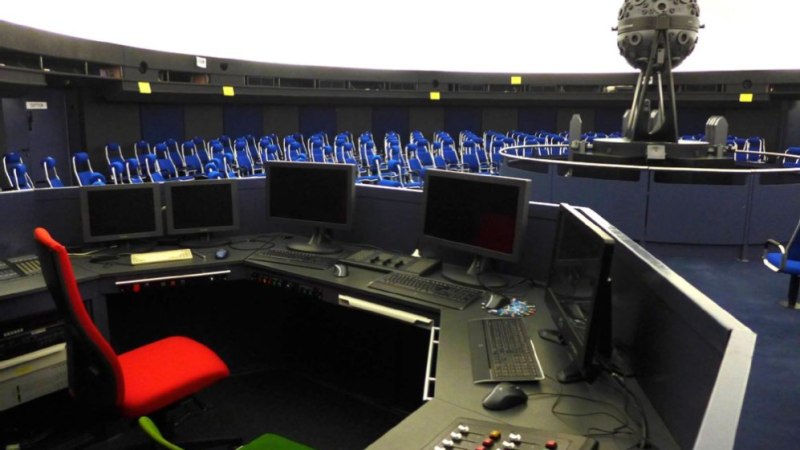 Kontrollpult mit Bildschirmen und Tasten in einem Planetarium. Im Hintergrund sind blaue Sitzreihen und ein großer Projektor zu sehen., © Planetarium Stuttgart Kontrollpult mit Bildschirmen und Tasten in einem Planetarium. Im Hintergrund sind blaue Sitzreihen und ein großer Projektor zu sehen., © Planetarium Stuttgart
