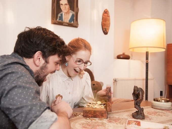 A man and a woman enjoy an open treasure chest full of coins in a cozy room., &copy; ExitGames Stuttgart