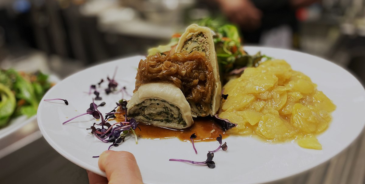 A plate of Swabian Maultaschen, garnished with onions and potato salad, is held in one hand. A kitchen can be seen in the background., © Speisekammer West