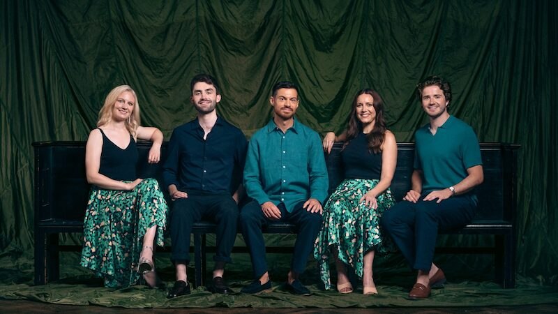 Fünf Personen sitzen lächelnd auf einer Bank vor einem grünen Vorhang. Sie tragen farblich abgestimmte Kleidung in Blau- und Grüntönen., © Stiftsmusik Stuttgart