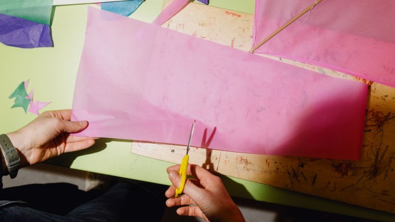 Eine Person schneidet rosa Papier mit einer gelben Schere. Auf dem Tisch liegen bunte Papierst&uuml;cke und ein Holzbrett., &copy; Linden-Museum Stuttgart, Foto: Harald V&ouml;lkl