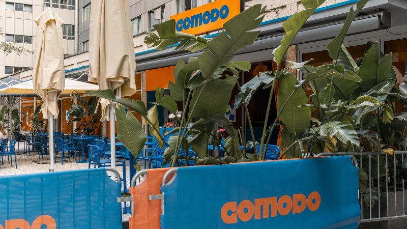 Au&szlig;enbereich eines Caf&eacute;s mit blauen St&uuml;hlen, Tischen und Pflanzen. Das Schild 'comodo' ist sichtbar. Sonnenschirme sind aufgestellt., &copy; Stuttgart Marketing GmbH, Sarah Schmid