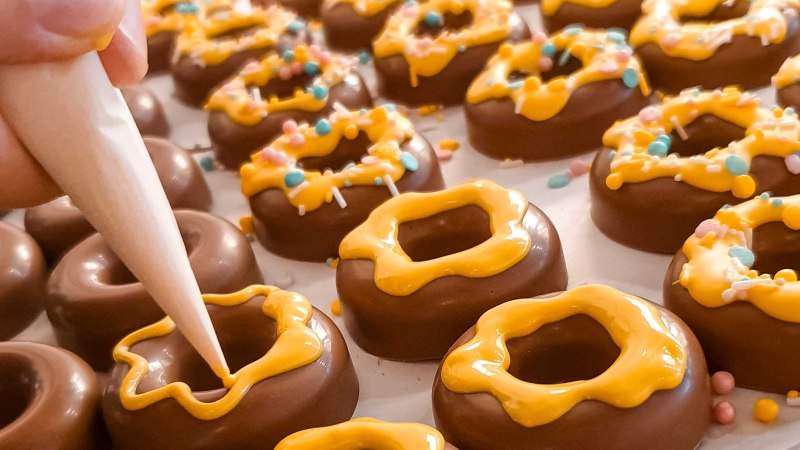 Schokoladenringe werden mit gelber Glasur und bunten Streuseln verziert. Eine Hand benutzt einen Spritzbeutel f&uuml;r die Dekoration., &copy; Kesselgl&uuml;ck
