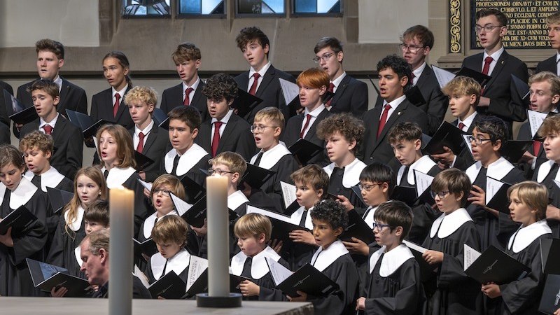 Ein Chor von Jungen und jungen M&auml;nnern singt in einer Kirche. Sie tragen schwarze Roben mit wei&szlig;en Kragen. Im Vordergrund stehen zwei brennende Kerzen., &copy; Stuttgarter Hymnus-Chorknaben