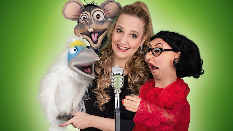 Eine Frau mit drei Puppen, darunter ein Vogel, eine Maus und eine Frau mit Brille, posiert fr&ouml;hlich vor einem Mikrofon auf gr&uuml;nem Hintergrund., &copy; Theaterhaus Stuttgart e.V.