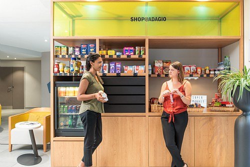 Two women are chatting in front of a small hotel store selling snacks and drinks. The store bears the sign 'SHOP@ADAGIO'., &copy; adagio access Stuttgart Airport Messe