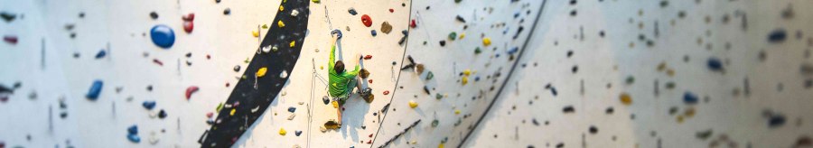 Ein Kletterer in grüner Kleidung erklimmt eine hohe Indoor-Kletterwand mit bunten Griffen im DAV Kletter- und Boulderzentrum Schwaben., © Rockerei