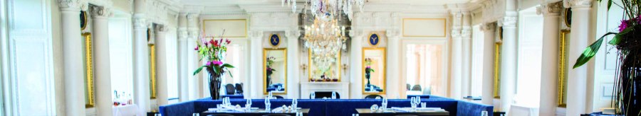 Elegant restaurant with chandelier, blue benches and white pillars. Flower arrangements and laid tables create a luxurious atmosphere., © Speisemeisterei