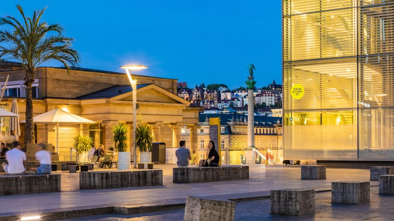 Evening atmosphere at Kleiner Schlossplatz with palm trees, people and modern architecture. Illuminated buildings and a statue can be seen in the background., &copy; SMG, Werner Dieterich