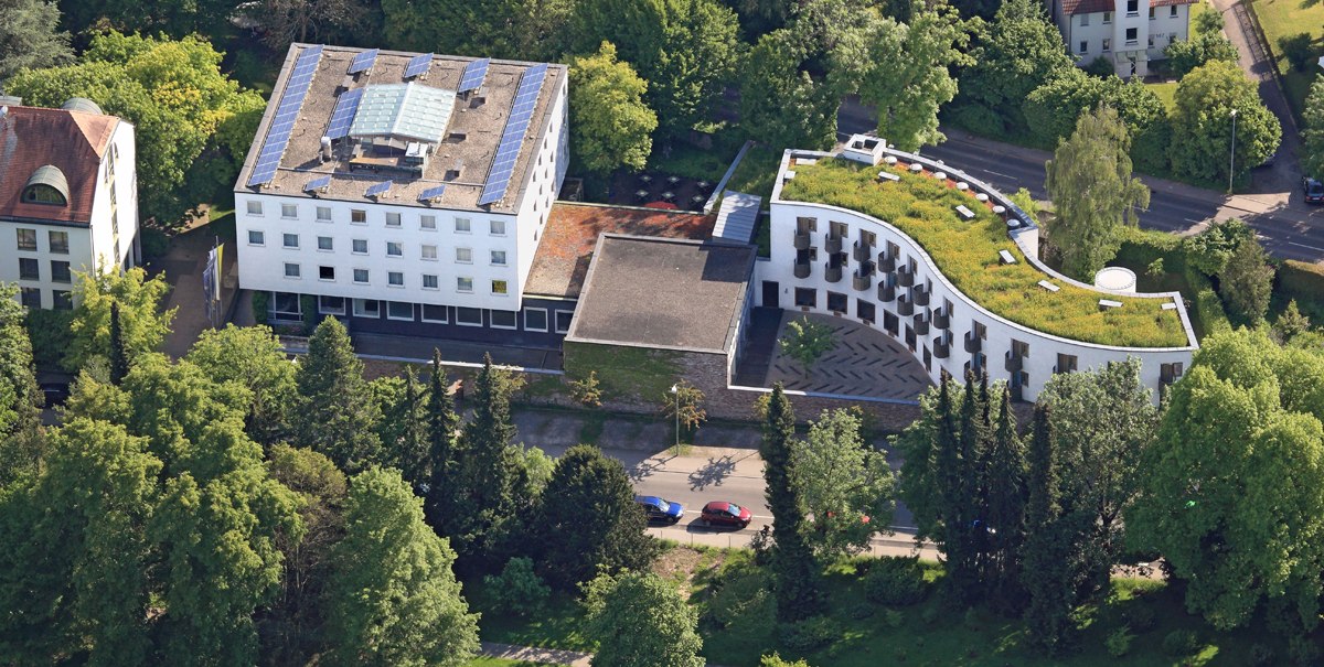 Luftaufnahme eines modernen Gebäudekomplexes mit begrüntem Dach, umgeben von Bäumen und Grünflächen. Solarpaneele sind auf dem Dach sichtbar., © TOMAS Luftaufnahme eines modernen Gebäudekomplexes mit begrüntem Dach, umgeben von Bäumen und Grünflächen. Solarpaneele sind auf dem Dach sichtbar., © TOMAS