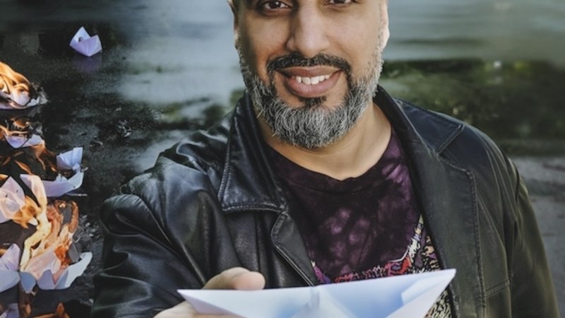 Ein Mann mit Glatze und Bart hält ein Papierboot. Im Hintergrund brennen weitere Boote auf einer nassen Straße., © Renitenztheater Stuttgart e.V. Ein Mann mit Glatze und Bart hält ein Papierboot. Im Hintergrund brennen weitere Boote auf einer nassen Straße., © Renitenztheater Stuttgart e.V.