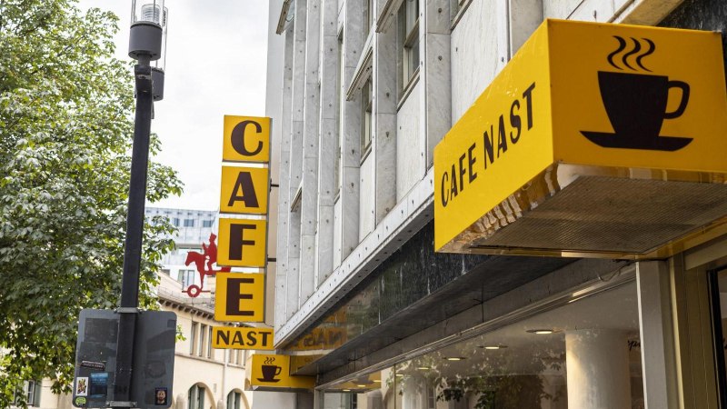 Gelbes Schild des Cafe Nast mit Kaffeetasse-Logo an einem Geb&auml;ude, umgeben von B&auml;umen., &copy; SMG, Sarah Schmid