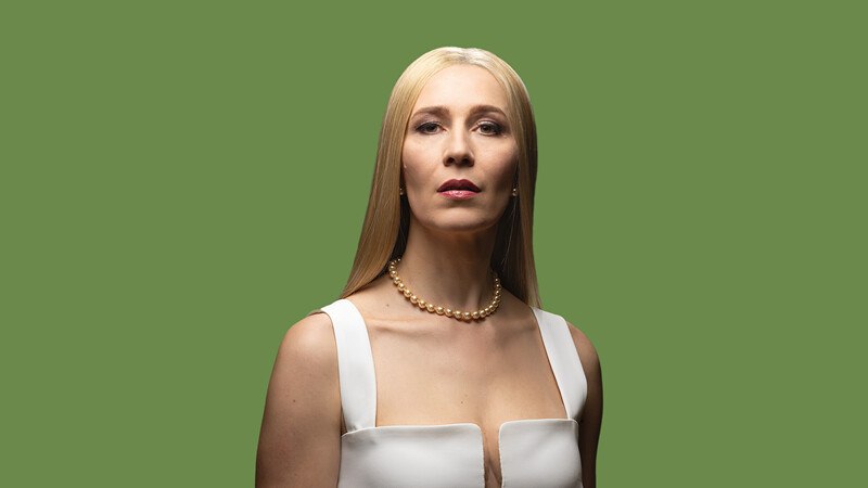 Eine Frau mit langen blonden Haaren trägt ein weißes Kleid und eine Perlenkette. Sie steht vor einem grünen Hintergrund und blickt selbstbewusst in die Kamera., © Württembergische Staatstheater Stuttgart Eine Frau mit langen blonden Haaren trägt ein weißes Kleid und eine Perlenkette. Sie steht vor einem grünen Hintergrund und blickt selbstbewusst in die Kamera., © Württembergische Staatstheater Stuttgart