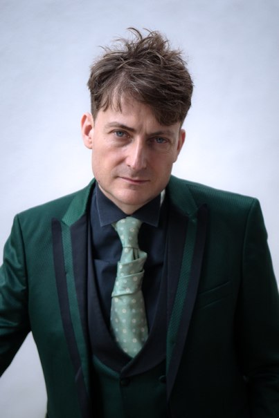 Ein Mann trägt einen eleganten grünen Anzug mit passender Krawatte und schaut direkt in die Kamera. Der Hintergrund ist schlicht und hell., © Rosenau Kultur e.V.