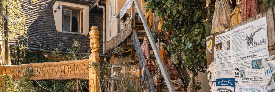 Eingang des Theater am Faden mit geschnitztem Holzschild, Puppen an einer Treppe und Plakaten an der Wand. Pflanzen umrahmen die Szene., &copy; SMG, Sarah Schmid