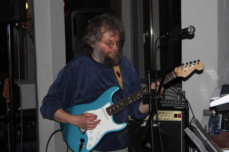 Ein Mann mit Brille und Bart spielt eine blaue E-Gitarre vor einem Mikrofon in einem Innenraum., © LonesomeRider