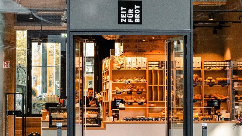 Eingang einer Bäckerei mit dem Schild 'Zeit für Brot'. Innen sind Regale mit Brot und Gebäck zu sehen., © Zeit für Brot Stuttgart
