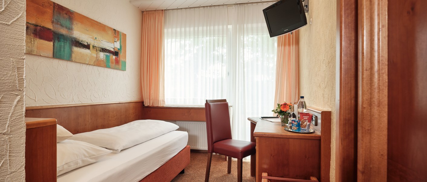 A cozy single room with a bed, desk, chair, and wall-mounted TV. Light-colored curtains and a modern painting adorn the room., © Badhotel Restaurant Stauferland A cozy single room with a bed, desk, chair, and wall-mounted TV. Light-colored curtains and a modern painting adorn the room., © Badhotel Restaurant Stauferland