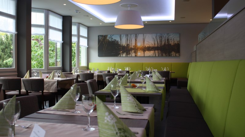 Modernes Restaurant mit gedeckten Tischen, grünen Servietten und großen Fenstern. An der Wand hängt ein Bild mit Sonnenuntergangsmotiv., © Hotel am Schlossberg Herrenberg Modernes Restaurant mit gedeckten Tischen, grünen Servietten und großen Fenstern. An der Wand hängt ein Bild mit Sonnenuntergangsmotiv., © Hotel am Schlossberg Herrenberg
