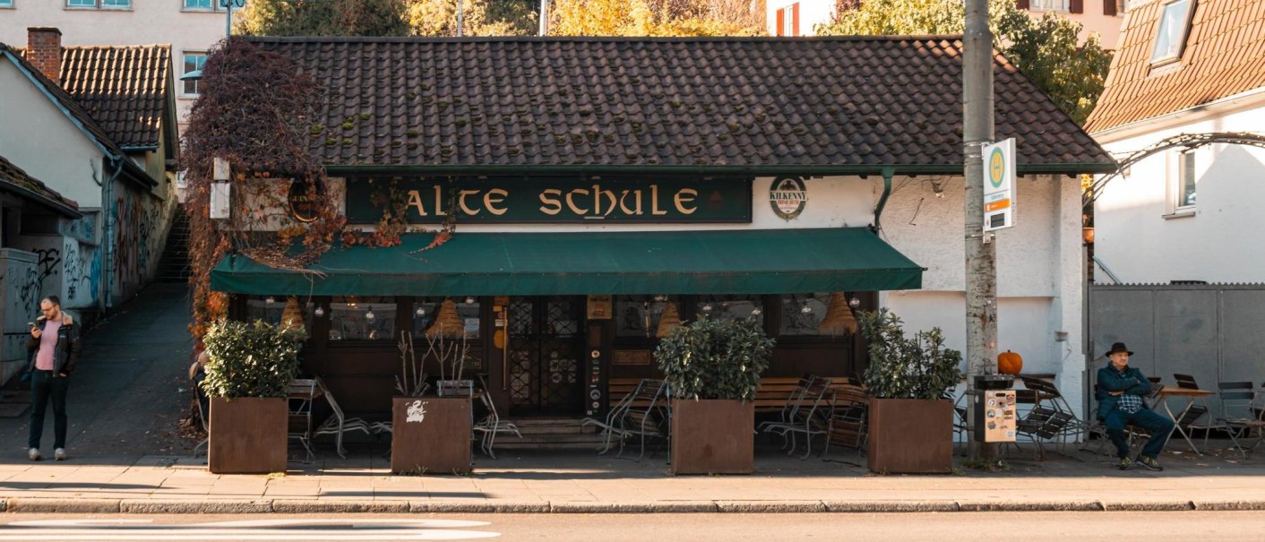 A cozy building with the sign 'Alte Schule' (old school), surrounded by plants. A man sits on a bench while another looks at his cell phone., &copy; SMG Stuttgart Marketing GmbH - Sarah Schmid