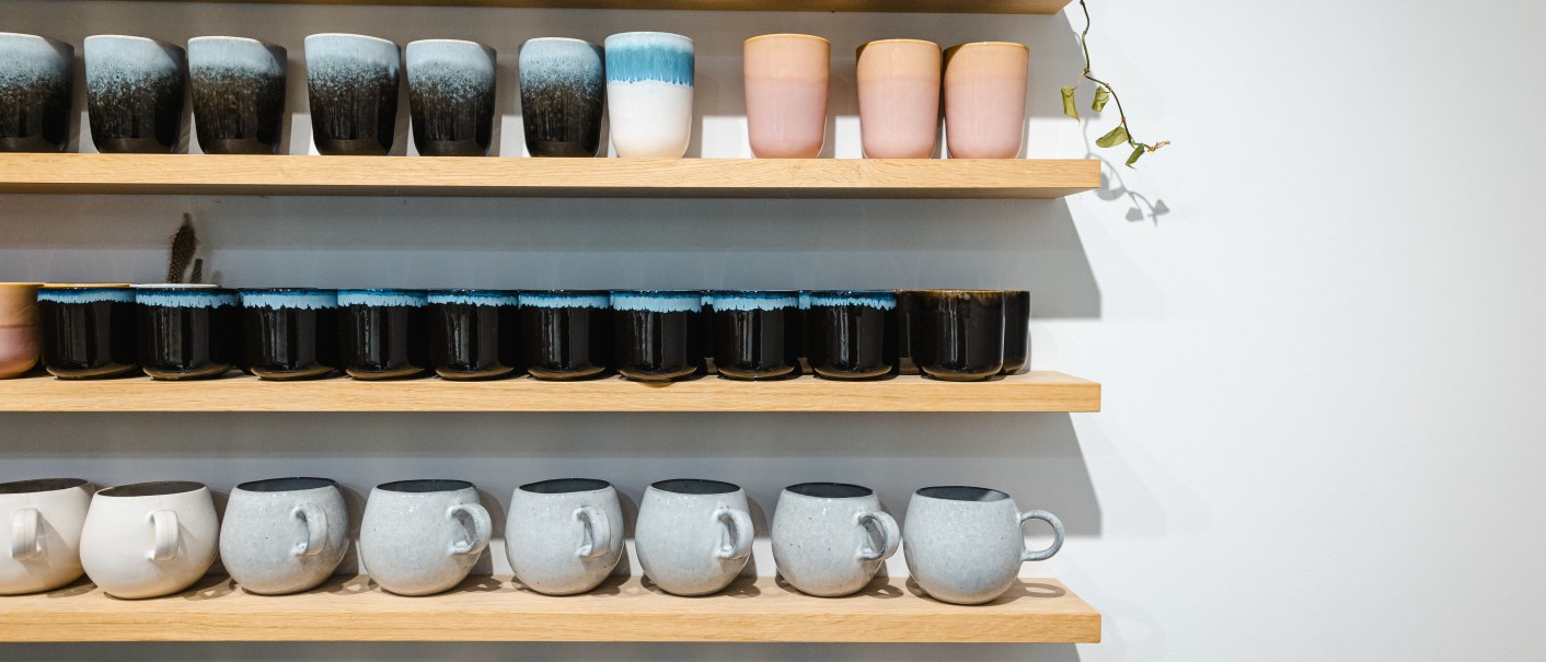 Holzregale mit bunten Keramiktassen und Bechern in verschiedenen Farben und Mustern. Eine Pflanze rankt am oberen Regal entlang., © SMG Stuttgart Marketing GmbH - Sarah Schmid Holzregale mit bunten Keramiktassen und Bechern in verschiedenen Farben und Mustern. Eine Pflanze rankt am oberen Regal entlang., © SMG Stuttgart Marketing GmbH - Sarah Schmid