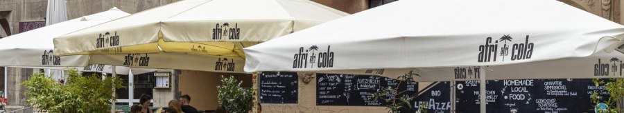 Außenbereich eines Cafés namens Platzhirsch mit Sonnenschirmen und Gästen. Menschen sitzen an Tischen und genießen Getränke., © SMG, Sarah Schmid