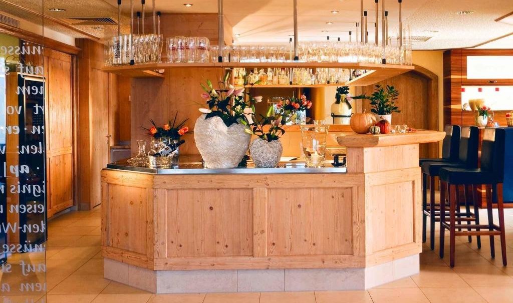 Cozy wooden bar with flower vases and glasses, surrounded by bar stools. The furnishings are inviting and warm., © Nödinger Gastro GmbH