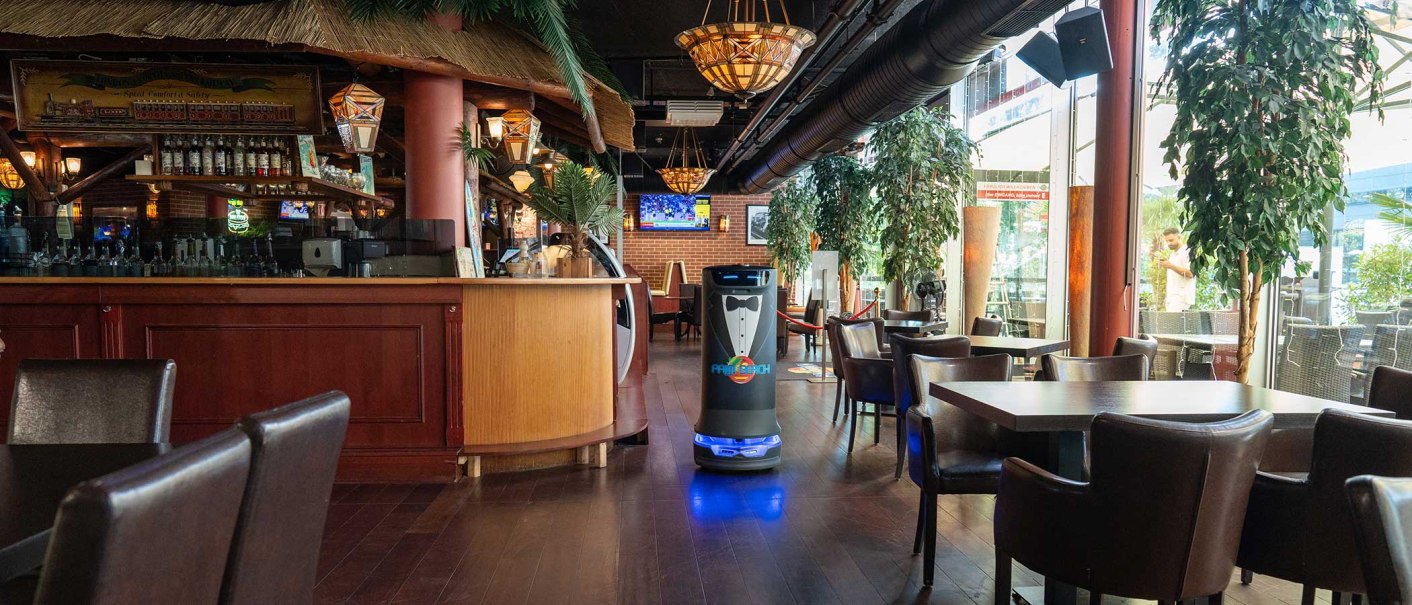 Ein Restaurant mit tropischer Dekoration, Holzmöbeln und einem Serviceroboter. Große Fenster lassen viel Licht herein, Pflanzen schmücken den Raum., © Palm Beach Stuttgart GmbH