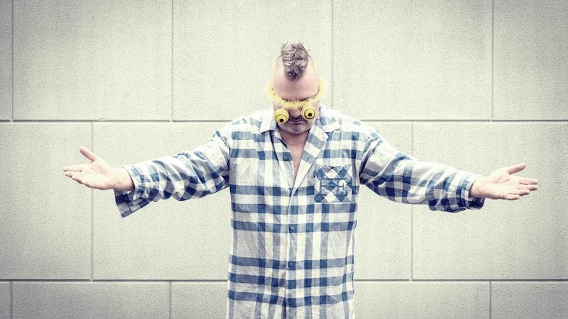 A person in blue and white checked pyjamas wears yellow glasses and stands with outstretched arms in front of a gray wall., &copy; Rosenau Kultur e.V.